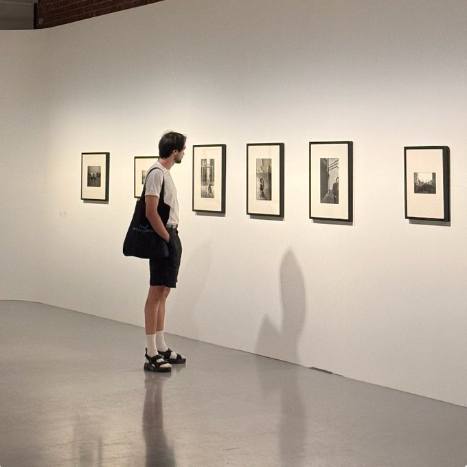 A person wearing a white t-shirt, black shorts, and sandals stands in a brightly lit art gallery, viewing a row of six framed photographs displayed on a white wall.