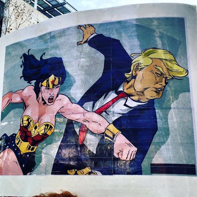 A poster or mural depicts Wonder Woman in her classic red, blue, and gold costume striking a dynamic fighting pose alongside a blonde man in a suit who is recoiling from her punch.