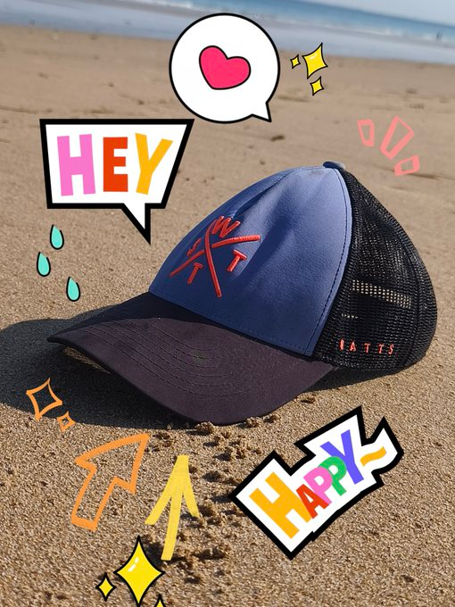A blue and black baseball cap on sandy beach ground, overlaid with cartoon-style speech bubbles and stickers reading 'HEY', 'HAPPY', and featuring a heart icon.