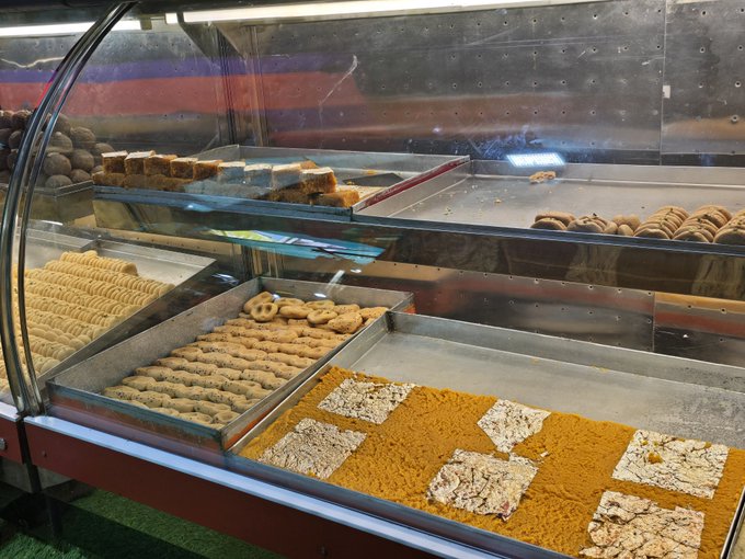 Photograph of a bakery display case filled with various pastries, cookies arranged in neat rows, pretzels, and decorative checkered patterns made from oats and golden-colored ingredients.