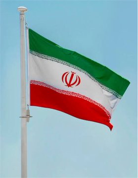 An Iranian flag (green, white, and red with the national emblem) waves on a flagpole against a clear blue sky.