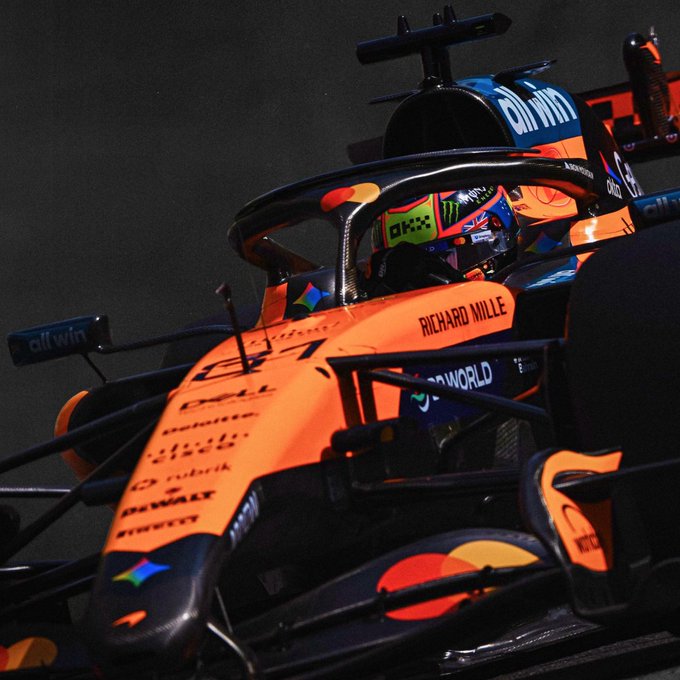 A close-up shot of an orange McLaren Formula 1 car showing the cockpit area, with the driver wearing a helmet featuring Monster Energy and OXS branding, and visible sponsors including "allwyn," "Richard Mille," and "Dell" on the chassis. The image captures the intense preparation moment, fitting the caption "Row two locked. Tomorrow is all throttle," as the driver readies for the next day's racing action.