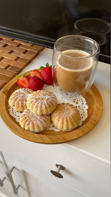 A wooden tray on a white dresser holding five powdered sugar cookies, fresh strawberries, and a double-walled glass mug of coffee or latte.