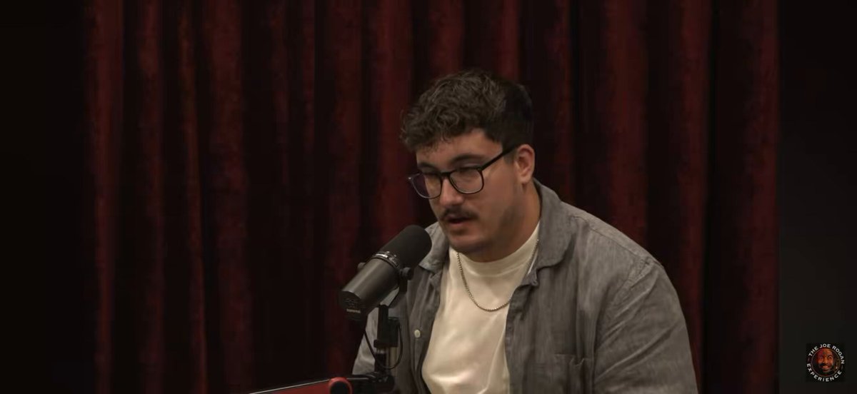 The image features a man wearing glasses, sitting at a microphone with his back towards the camera. He appears to be speaking into a microphone or giving a speech. The background of the image shows a red curtain, which could potentially be part of a stage setting for an event or performance. There is also text in the image that reads "How kaspa people sound: 
Guys ths is the new Bitcoin!! It' s called hawkTuah and it was made by israeli intelligence". This suggests that there might be some sort of discussion or presentation related to cryptocurrency, possibly involving the man speaking into a microphone.