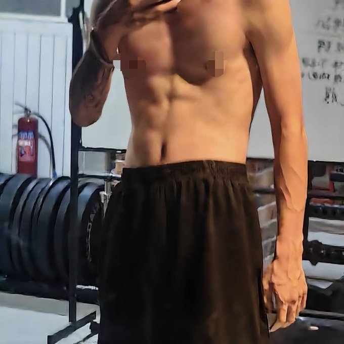 A shirtless man in a gym poses to showcase his defined abs and muscular arms, with a fire extinguisher and weight plates visible in the background. The image illustrates his five-month fitness transformation, with the visible muscle definition reflecting the early results of his training progress.