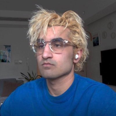 A man with messy blonde hair, clear-framed glasses, and a white earbud wears a blue sweatshirt in an indoor setting.