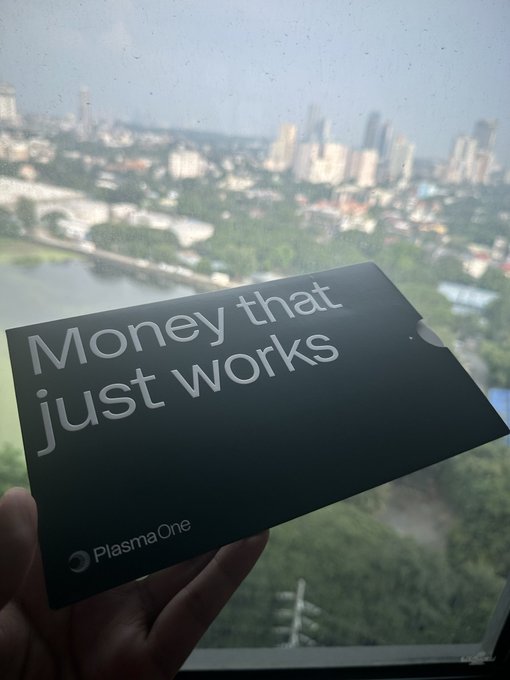 A hand holding a black card with the text 'Money that just works' and the 'PlasmaOne' logo, with a city skyline visible through a window in the background.