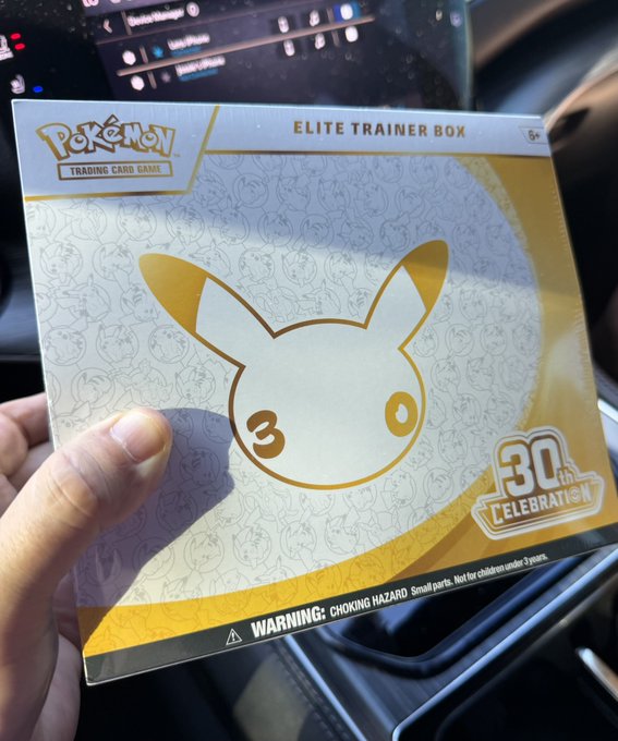 A hand holds a Pokemon Trading Card Game Elite Trainer Box featuring Pikachu in gold and white, commemorating the 30th anniversary celebration, photographed inside a vehicle.