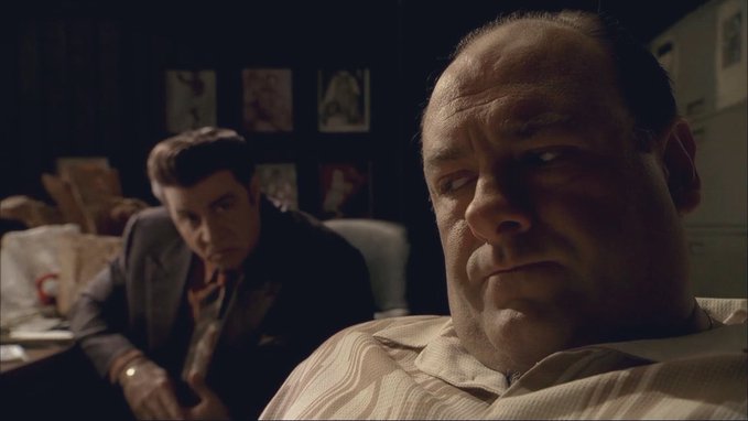 A dimly lit scene from a TV show featuring two men in a dark room, with a balding man in the foreground and a younger dark-haired man leaning in the background, with framed pictures visible on the wall.