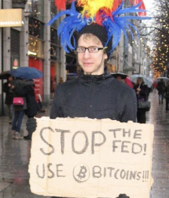 A young man wearing a colorful feathered headdress and glasses stands on a rainy street holding a handwritten cardboard sign that reads "STOP THE FED! USE BITCOINS!!!" with the Bitcoin "₿" symbol, illustrating the post's reference to someone advocating Bitcoin when its price was just $0.30.