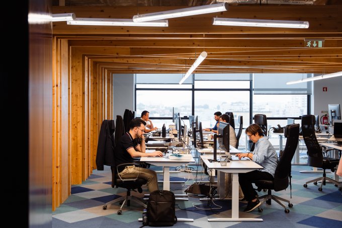 A modern, open-plan office space with wooden beam ceilings and large windows shows several team members working at their desks on computers, fitting the "Let's build!" theme of a productive and collaborative September kickoff. An emergency exit sign is visible in the upper right corner of the room.