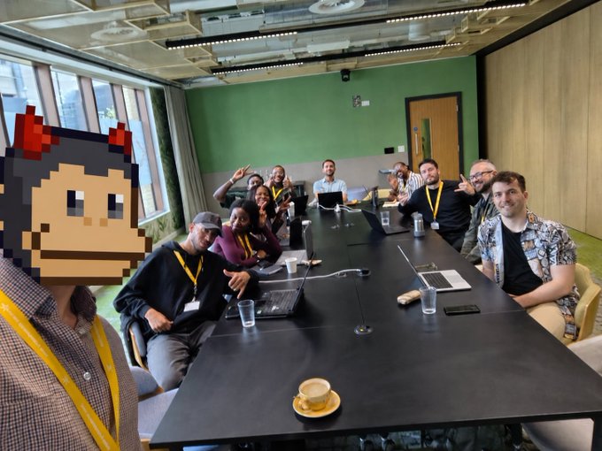 A group of people wearing yellow lanyards pose around a long conference table in a meeting room, with one person in the foreground wearing a pixel art cat mask.