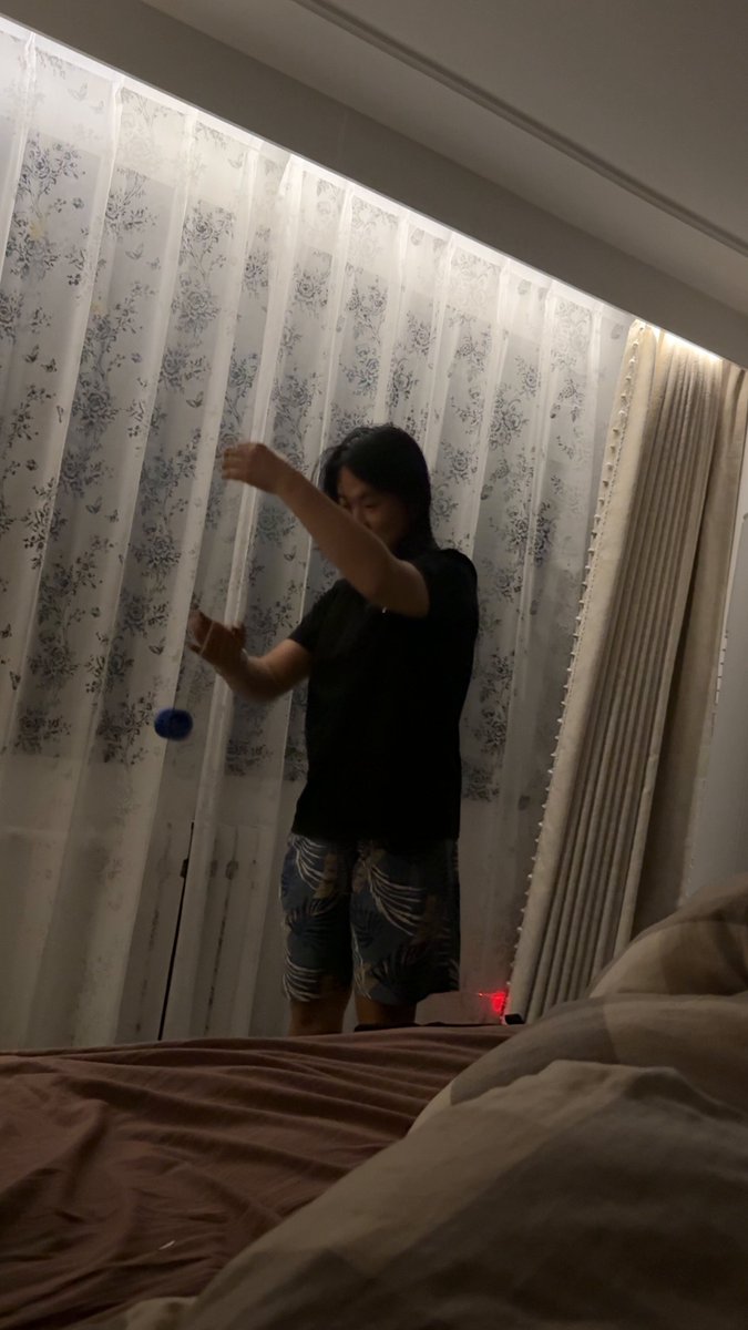 The image shows a person standing in front of a window wearing shorts and a black shirt. They are playing with a toy, possibly a yo-yo or a ball, which they are holding up against the backdrop of the window. The room appears to be a bedroom, as there is a bed visible in the background.