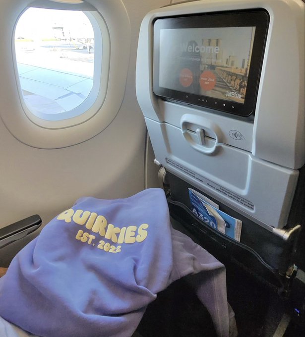 A photo taken inside an airplane showing a JetBlue seatback screen displaying 'Welcome' and a 'Quirkies EST. 2025' sweatshirt resting on the tray table.