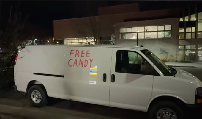 A white cargo van parked at night with 'FREE CANDY' spray-painted in red on its side panel, in front of an office building.