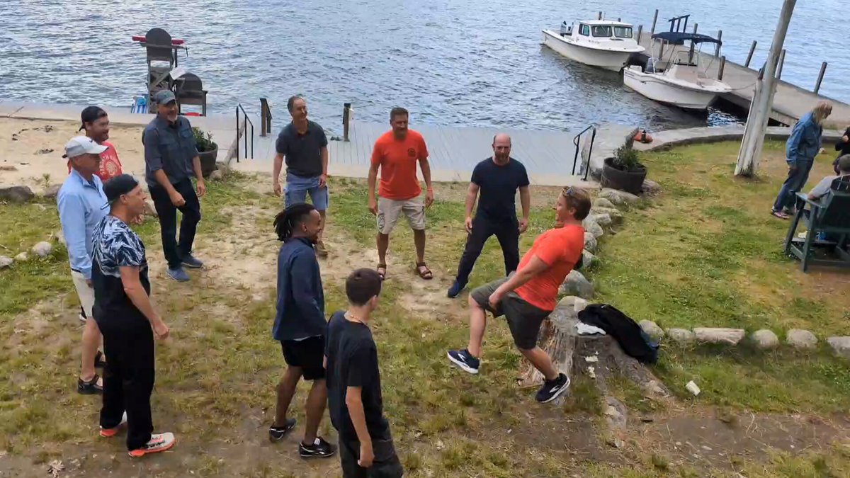 The image shows a group of people standing around, with some playing frisbee near a body of water. There are at least nine people visible in the scene, including two men who appear to be engaged in a friendly competition or duel over the frisbee. The presence of boats and a dock suggests that this gathering might be taking place on a lake or riverfront location.