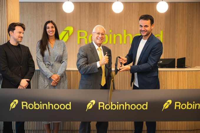 Four people stand behind a black ribbon at the opening of Robinhood's new Toronto office, with two men in the center using scissors to cut the ribbon while two others smile beside them. The "Robinhood" logo is prominently displayed on both the ribbon and the reception desk backdrop, marking the company's expanded Canadian presence.
