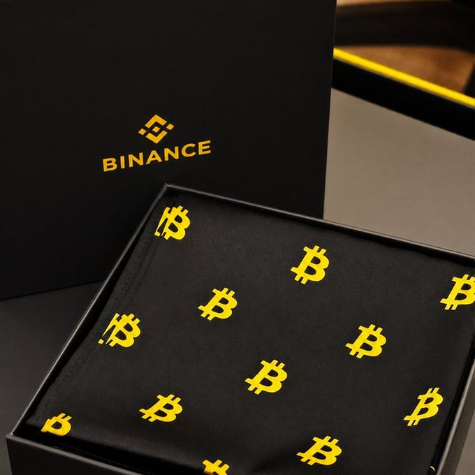 A black gift box displays the 'BINANCE' logo, containing a folded cloth item patterned with yellow Bitcoin (₿) symbols.