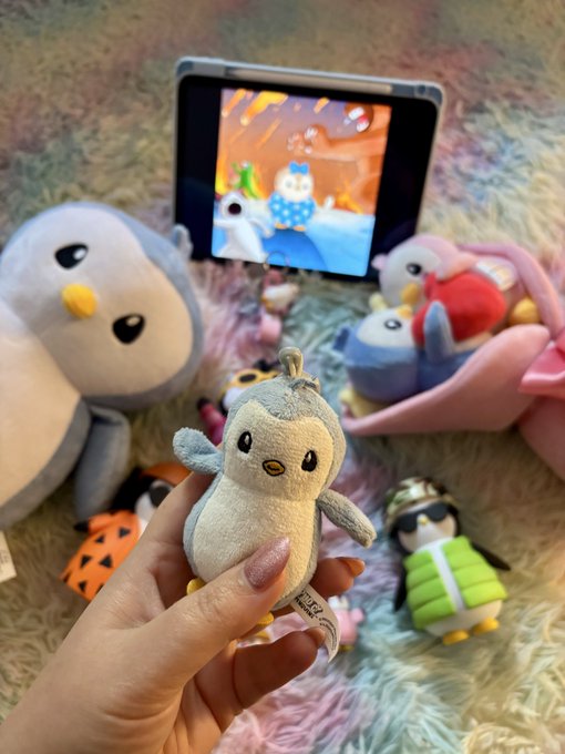 A hand holds up a small gray and cream penguin plush toy amid a cozy collection of stuffed animals and other penguin figures, with a tablet playing what appears to be a Pudgy Penguins animation in the background. The image perfectly illustrates the poster's devotion to the Pudgy Penguins NFT brand, showcasing a heartfelt display of penguin-themed merchandise.