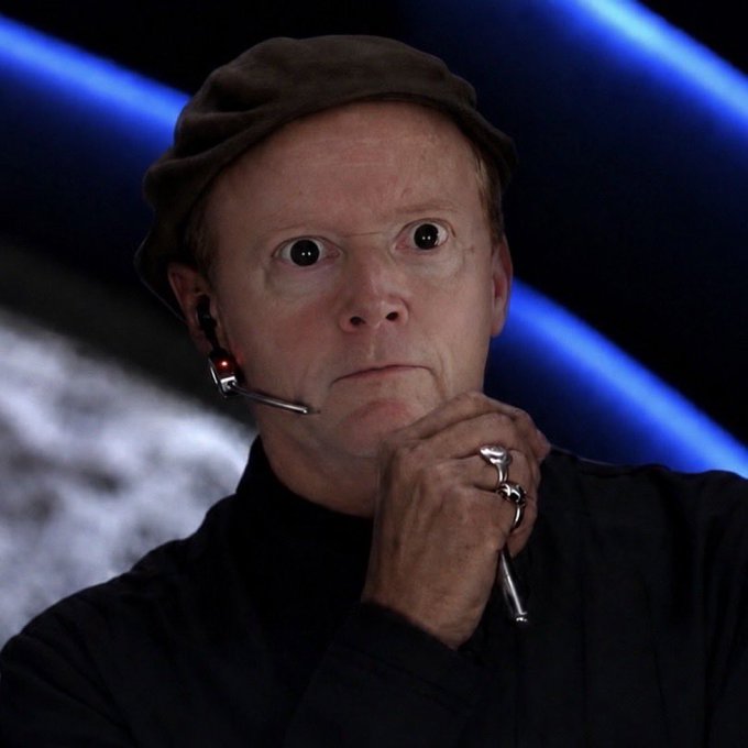 A man wearing a black shirt and brown cap, with wide eyes and a surprised expression, resting his chin on his hands, with silver skull rings on his fingers and a headset microphone.
