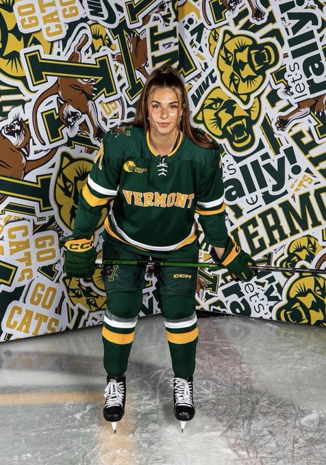 Hilary Wilkin of Vermont stands on the ice in the Catamounts' green and gold uniform, posing with her stick against a backdrop of Vermont-themed graphics featuring "GO CATS" and bear mascot logos.