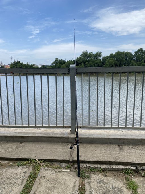 A fishing rod stands upright leaning against a metal railing beside a calm river under a partly cloudy sky.
