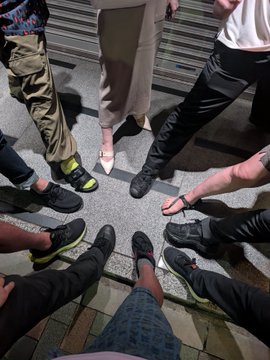 A low-angle shot of a group of people standing in a circle on outdoor stairs, showing only their legs and shoes, with one person holding a phone.