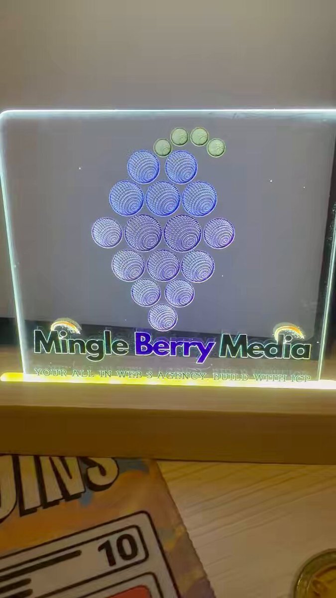 The image features a small glass display case containing a sign that says "Mingle Berry Media". The display case has a unique design, resembling a bunch of grapes or berries. It's placed on top of a wooden table, which adds a rustic touch to the overall presentation. In addition to the display case and its contents, there is also an image of a coin in the bottom right corner of the frame.