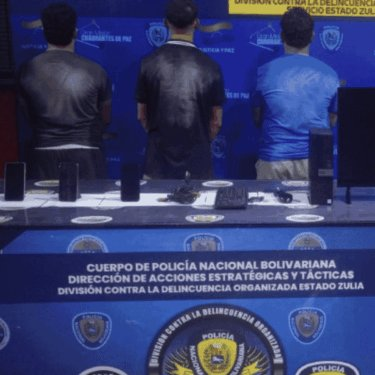 Three suspects stand facing a backdrop during a press conference by Venezuela's Cuerpo de Policía Nacional Bolivariana, with seized phones, laptops, and firearms displayed on a table in front of them. Visible text identifies the División Contra la Delincuencia Organizada Estado Zulia, marking the arrest of "Los Binanceros," a group accused of arbitraging the official Venezuelan dollar against USDT via Binance P2P.