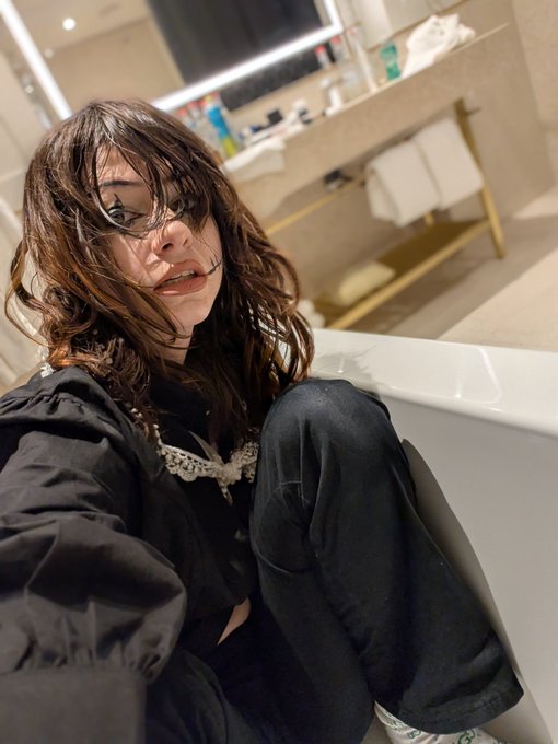iced tea and a green bottle are on the bathroom counter, with a woman in black clothing seated on the edge of a bathtub.