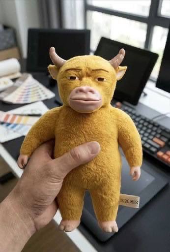 A hand holds a yellow plush cow toy with small horns and big lips in front of a desk with a keyboard and color swatches, matching the post's "Niu Bubu has arrived" as the cuddly new arrival.