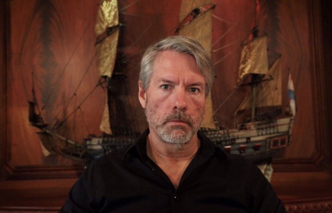A man with gray hair and a black shirt stands in front of a painting of a tall ship, his serious expression complementing the post's reflective tone about Michael Saylor and his long-term perspective on capital markets and technology.
