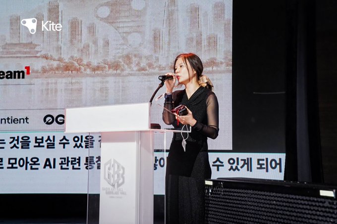 A woman in a black outfit with a red headband speaks into a microphone at a podium during a presentation, with a 'Kite' logo visible on the screen behind her.