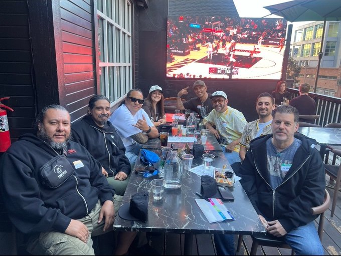 A group of friends gathers around an outdoor restaurant table beneath a TV broadcasting a basketball game, posing together as a close-knit "BAYC (Bored Ape Yacht Club) family." Their relaxed smiles and shared table of drinks and food reflect the camaraderie described in the caption.