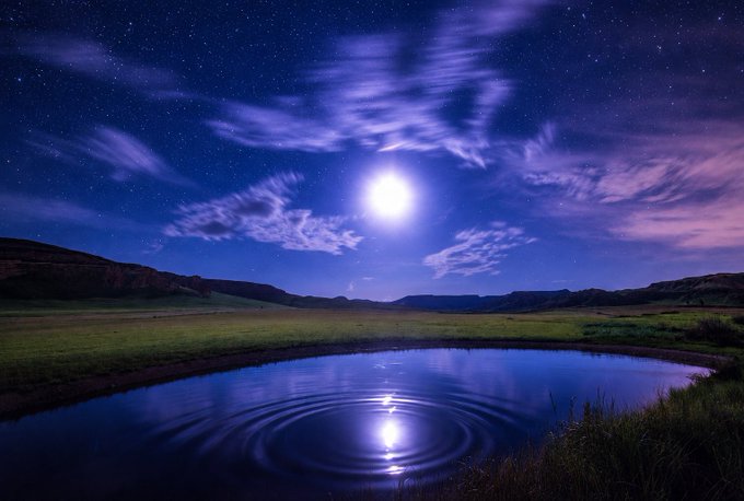 A serene night landscape featuring a bright full moon, starry purple-blue sky, clouds, and its reflection shimmering on a calm pond surrounded by grassy hills.