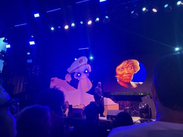 A concert stage illuminated in blue lighting features a large screen displaying an animated cowboy-hat-wearing character alongside a face, with a performer visible on stage and audience members' silhouettes in the foreground.