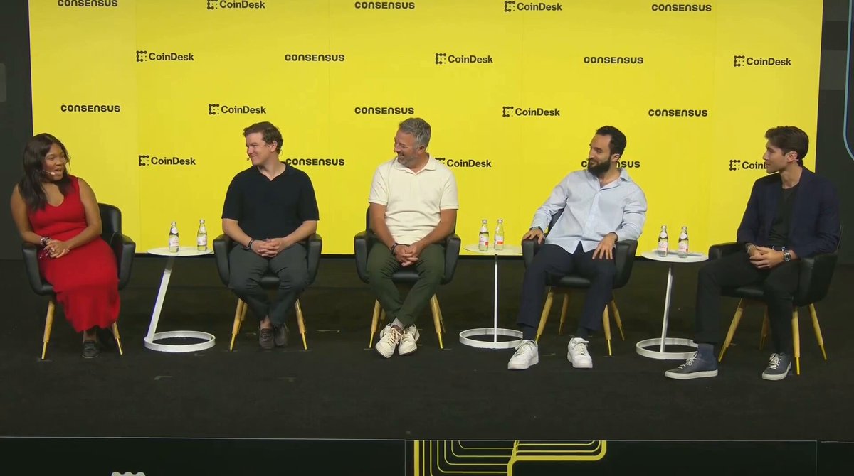 The image shows a group of five people sitting around a table, likely participating in a panel discussion or interview. They are all dressed professionally, with one person wearing a red dress. The backdrop features a yellow wall adorned with various logos, including "SoFi" and "Cards". The text on the wall reads: "What I'm really excited about is who's using it and why." This suggests that the panel discussion might be related to the SoFi partnership or the concept of cryptocurrency adoption.