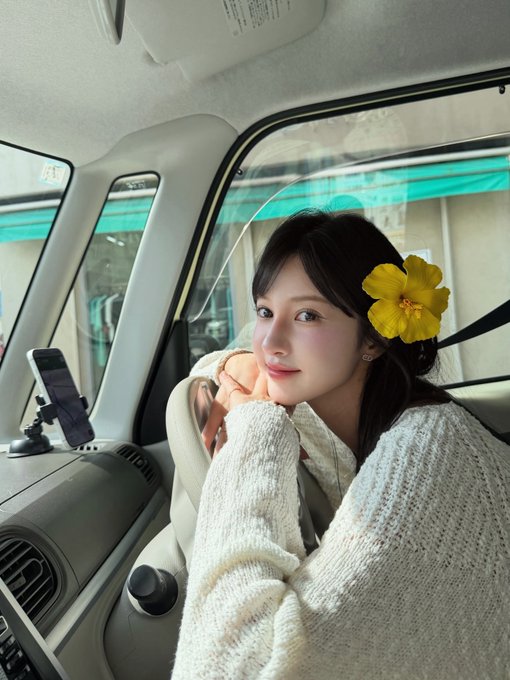 A young woman sitting in the passenger seat of a car, wearing a white knit cardigan with a yellow hibiscus flower tucked behind her ear.