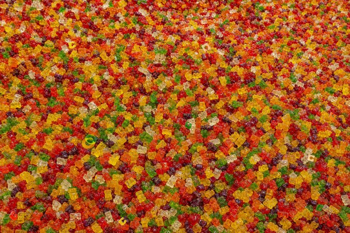 A close-up photograph of colorful gummy bear candies in red, orange, yellow, green, and clear/purple colors densely packed together.