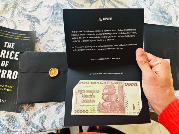 A hand displays a 'River' branded envelope containing a real Zimbabwean hyperinflation banknote (200,000,000 dollars) with a gold Bitcoin coin seal, promoting Bitcoin investment as protection against inflation.