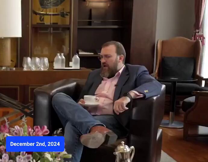The image shows a man sitting comfortably in a chair, holding a cup of coffee while wearing glasses and a suit. The room has wooden floors and furniture, including chairs and a table with flowers on it. There are also wine glasses and bottles present in the scene. A blue text box is visible in the bottom left corner of the image, which appears to be related to the Cardano Bitcoin DeFi thesis posted by Charles Hoskinson back in December 2024.
