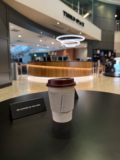A takeaway coffee cup with a sleeve sits on a black table next to a 'NO LAPTOPS IN THIS AREA' sign, with a 'THIRD WAVE' coffee counter visible in the blurred background of what appears to be a shopping mall.