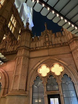 A nighttime photograph of an ornate Gothic-style stone building with a large rose window featuring a four-leaf clover design, illuminated by string lights.