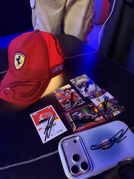 A red Ferrari Racing baseball cap featuring the prancing horse logo and Puma branding sits on a black surface alongside signed Formula 1 trading cards and a smartphone case.