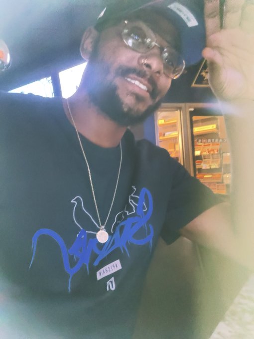 A selfie of a smiling man with glasses and a goatee wearing a dark blue cap and a black t-shirt with blue text, holding a pendant necklace.