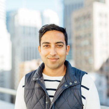 A man with short dark hair smiles at the camera, wearing a navy quilted vest over a striped shirt, with blurred city skyscrapers in the background. The image likely accompanies the post featuring Asheesh Birla, the Evernorth CEO discussing XRPL's role in payments.
