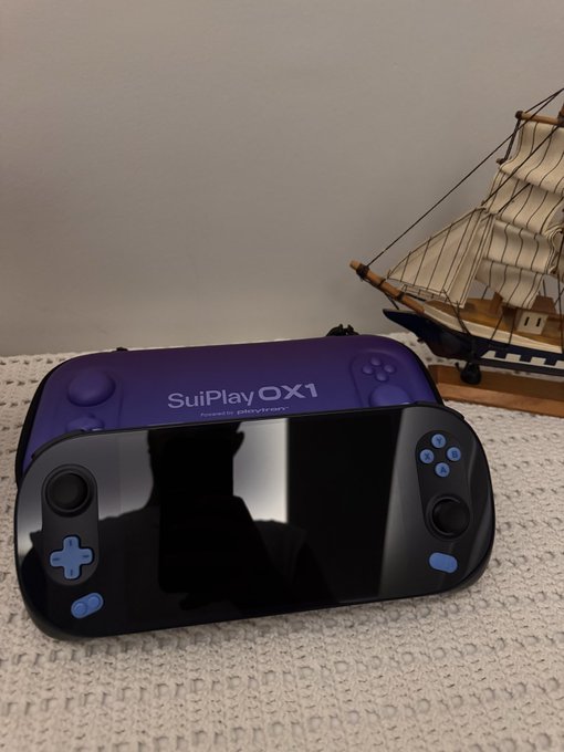 A purple SuiPlay0X1 handheld gaming console sits on a white knit surface next to a model sailboat. The device features the "SuiPlay0X1" branding and "Powered by playtron" text, along with blue accent buttons and a touchscreen display. This matches the post, as the user has received three of these SuiPlay handheld gaming devices they ordered a year ago, which have now arrived at their Switzerland home.