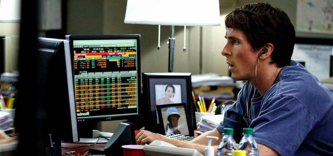 The image features an actor wearing headphones sitting behind a desk with two computer monitors and several keyboards, likely working or studying. The text displayed on his screens appears to be related to cryptocurrency trading. The scene suggests that he may be involved in the cryptocurrency market, where various virtual currencies like Bitcoin are being monitored for trading purposes.