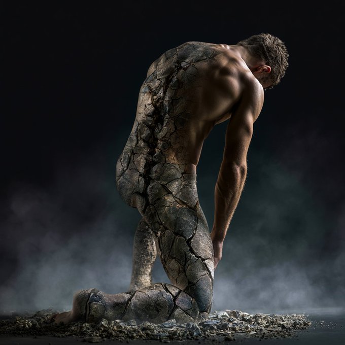A conceptual photo of a man kneeling in a dark, smoky environment, with his lower body appearing to be made of cracked stone or earth.