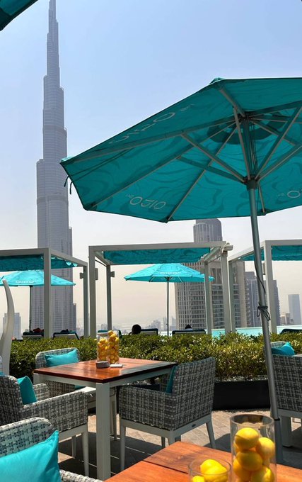An outdoor terrace restaurant with teal umbrellas and wicker chairs offers a view of the Burj Khalifa and Dubai skyline on a hazy day, with a glass of lemonade on the table.